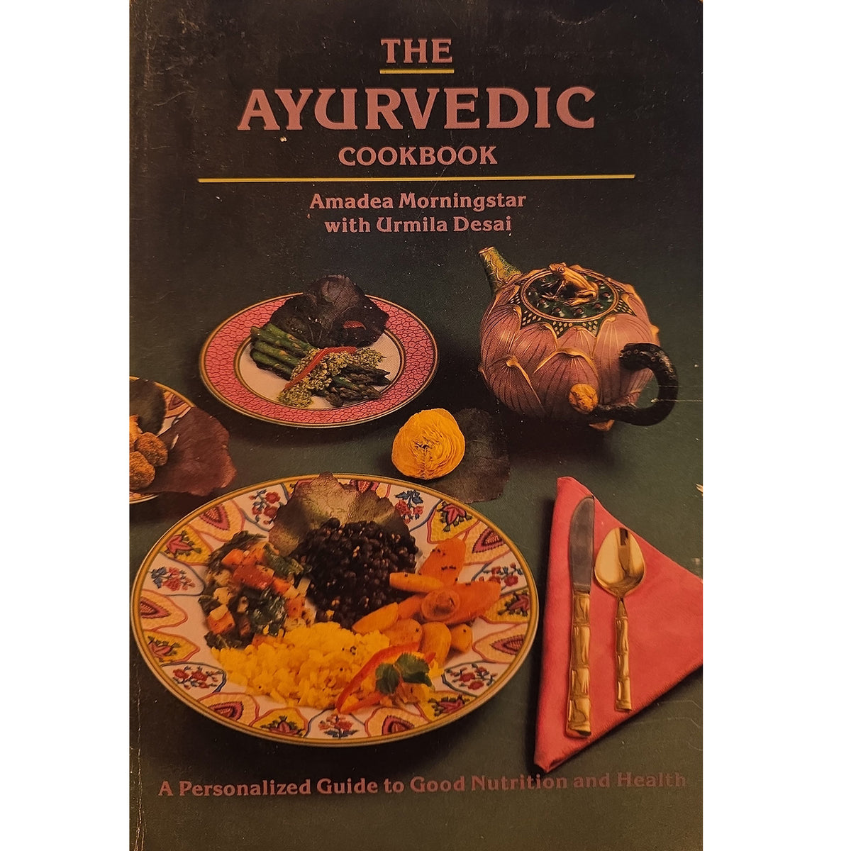 The Ayurvedic Cookbook by Amadea Morningstar with Urmila Desai, featuring a dark cover with traditional Indian dishes, a teapot, and a plate on a dark background.