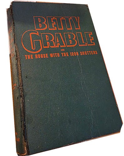 Original green cloth hardcover with orange lettering on spine, “Betty Grable and the House With the Iron Shutters,” showing vintage wear.
