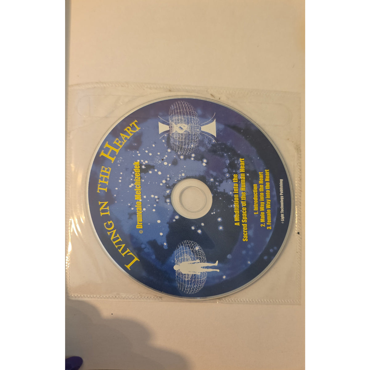 A blue compact disc with yellow text reading 'Living in the Heart' and whale graphics, placed on a white surface inside a clear plastic sleeve.