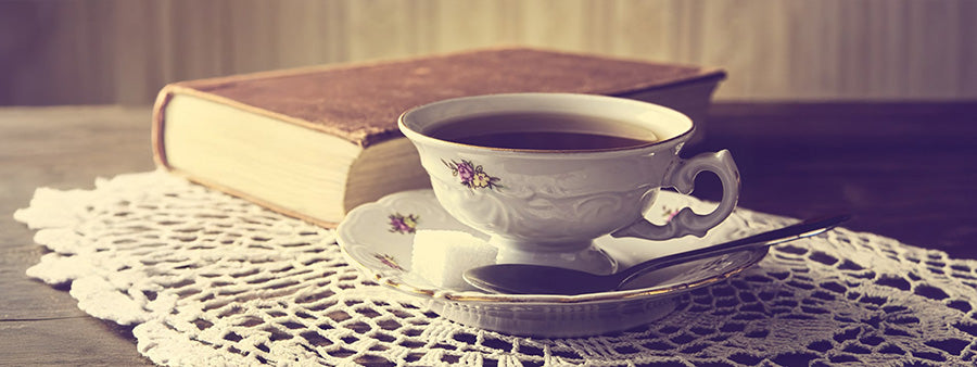 Vintage teacup and saucer with a sugar cube beside an old book on a lace doily — Deb’s Tea Room collection celebrating timeless comfort and literary charm.
