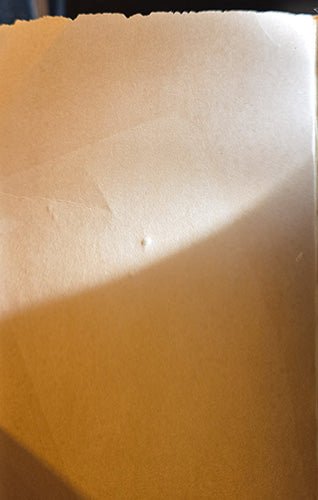 A close-up of a textured light brown kraft paper surface with a warm yellowish cast and a soft shadow across the lower-left area.