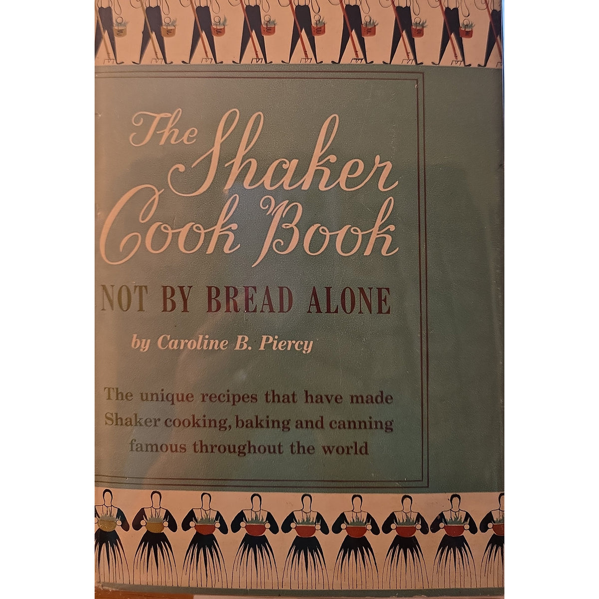 The Shaker Cook Book: Not By Bread Alone by Caroline B. Piereny, featuring a muted green cover with elegant script title text and decorative illustrations of Shaker figures at the top and bottom.