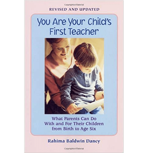 The book cover of You Are Your Child’s First Teacher features a warm image of a smiling parent guiding a young child who is reading. The text is in blue on a soft pastel pink background.