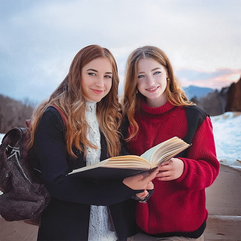 Two young women standing outdoors in winter, smiling while reading an open book together.