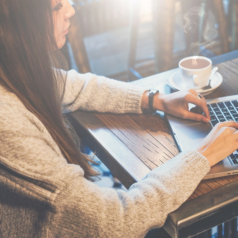 A woman in a cozy gray sweater types on a laptop at a wooden café table with a steaming cup of coffee beside her. Sunlight streams through the window, creating a warm and productive atmosphere.