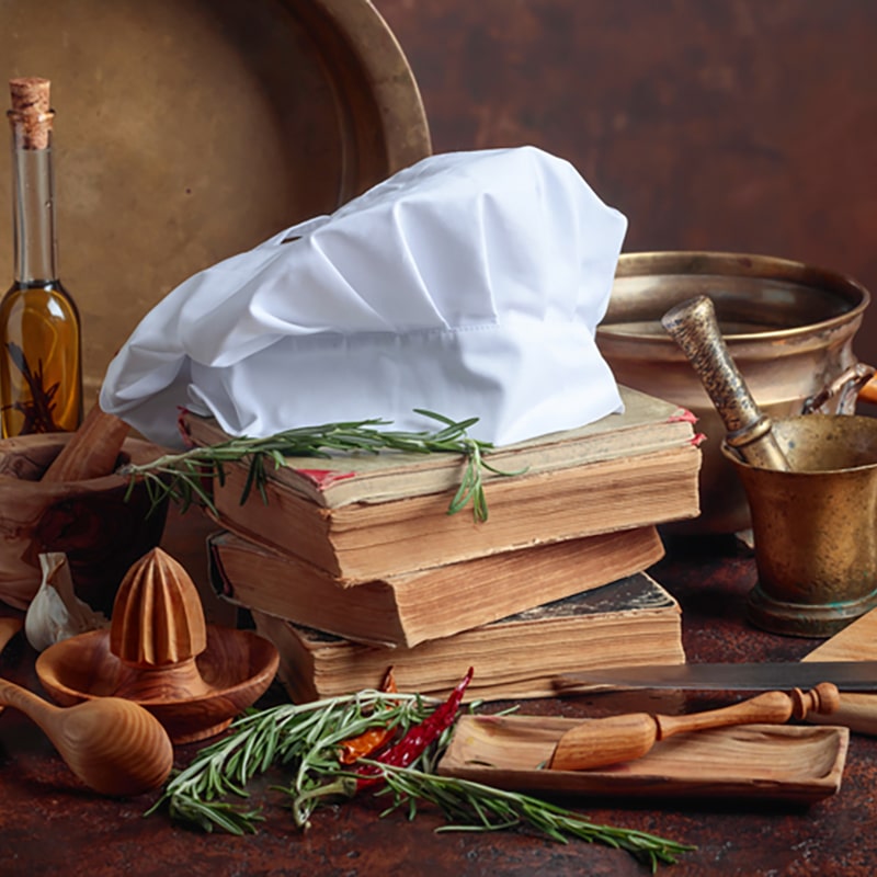 A stack of vintage cookbooks with worn pages sits on a rustic wooden table, topped with a white chef’s hat. Surrounding the books are fresh herbs, wooden utensils, an olive oil bottle, and brass kitchenware, creating a warm and nostalgic culinary scene.