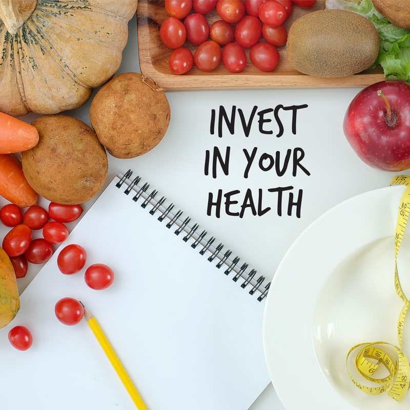 A flat lay of fresh vegetables, fruits, and a spiral notebook with a yellow pencil, set against a white background with the phrase "Invest in Your Health" written in bold. A white plate with a measuring tape symbolizes healthy eating and wellness.