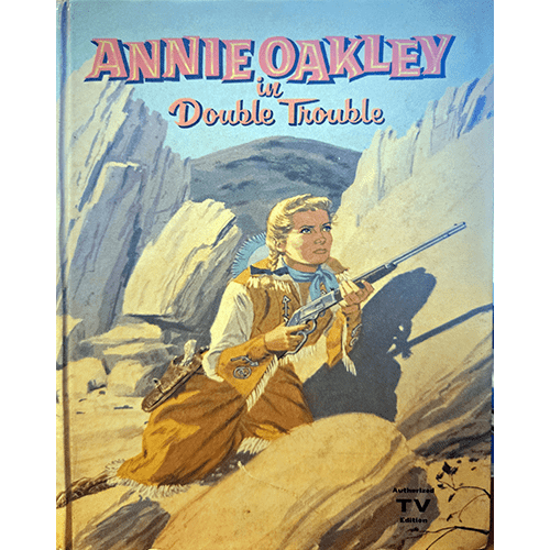 The cover of Annie Oakley in Double Trouble shows a vintage illustration of Annie Oakley in fringed buckskin, holding a rifle, set against rugged desert cliffs. A timeless nod to classic Western adventures.