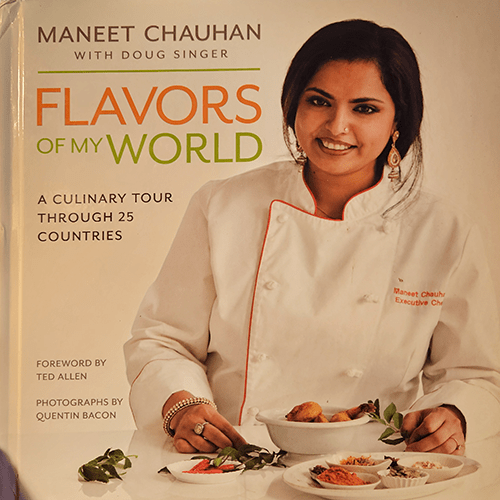 "Cover of Flavors of My World by Maneet Chauhan featuring the chef in a white coat with vibrant dishes in front. The book promises a culinary tour through 25 countries, blending flavors globally."