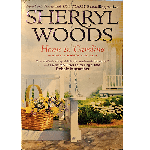 The cover of Home in Carolina by Sherryl Woods shows a peaceful porch scene with hanging flower baskets and a white wooden swing, evoking the warm, welcoming atmosphere of a small southern town.