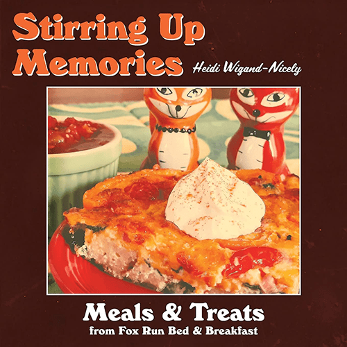 The book cover titled "Stirring Up Memories" by Heidi Wigand-Nicely features a photo of a savory dish topped with a dollop of cream. Behind the dish are two fox figurines. The title is in large orange letters, with "Meals & Treats from Fox Run Bed & Breakfast" below. The background is dark brown.