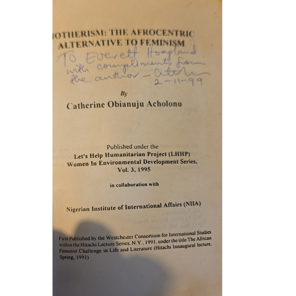 A close-up of a book cover or printed page with visible text including the title 'Motherhood: The Afrocentric Alternative to Feminism' and the author name Catherine Obianuju Achiolonu, plus a handwritten note in blue ink. The page is beige/tan with black text and a small amount of blue writing.