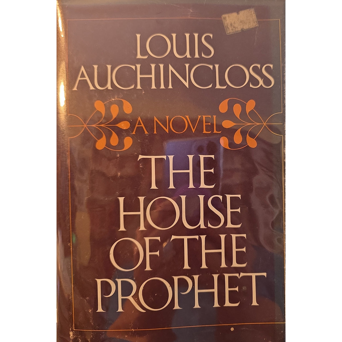 A vintage hardcover book of Louis Auchincloss's 'The House of the Prophet' with a glossy, reflective cover. The cover is dark brown with gold and orange lettering, and shows some wear, scuffs, and a small sticker near the top.