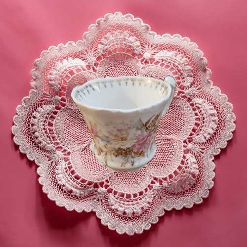 White teacup with floral design on a lace doily against a pink background