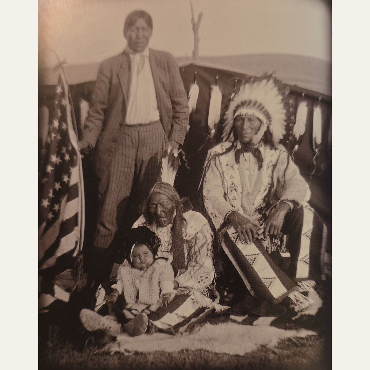 A sepia-toned vintage photograph showing three Native American individuals posed outdoors in front of a tent, with an American flag visible on the left. The scene is framed by a light border around the print.